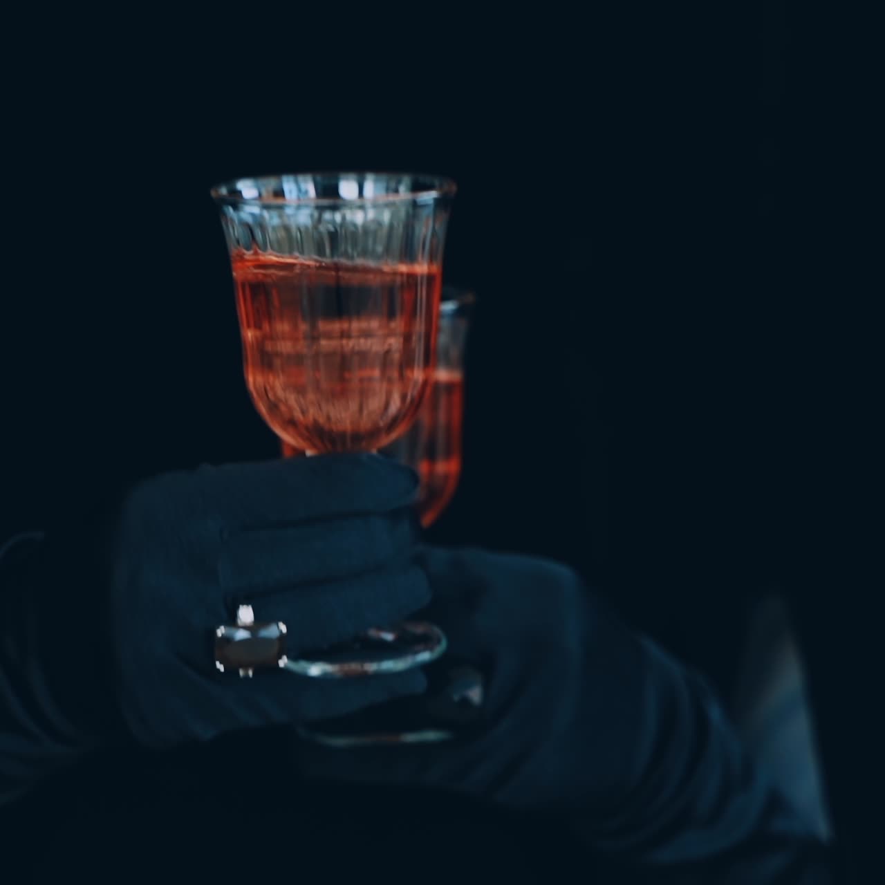 Two glasses of red champagne on the dark background. Enigmatic hands in luxury black gloves hold two glasses of wine.