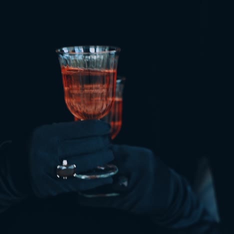 two glasses of red champagne on the dark background. enigmatic hands in luxury black gloves hold two glasses of wine.