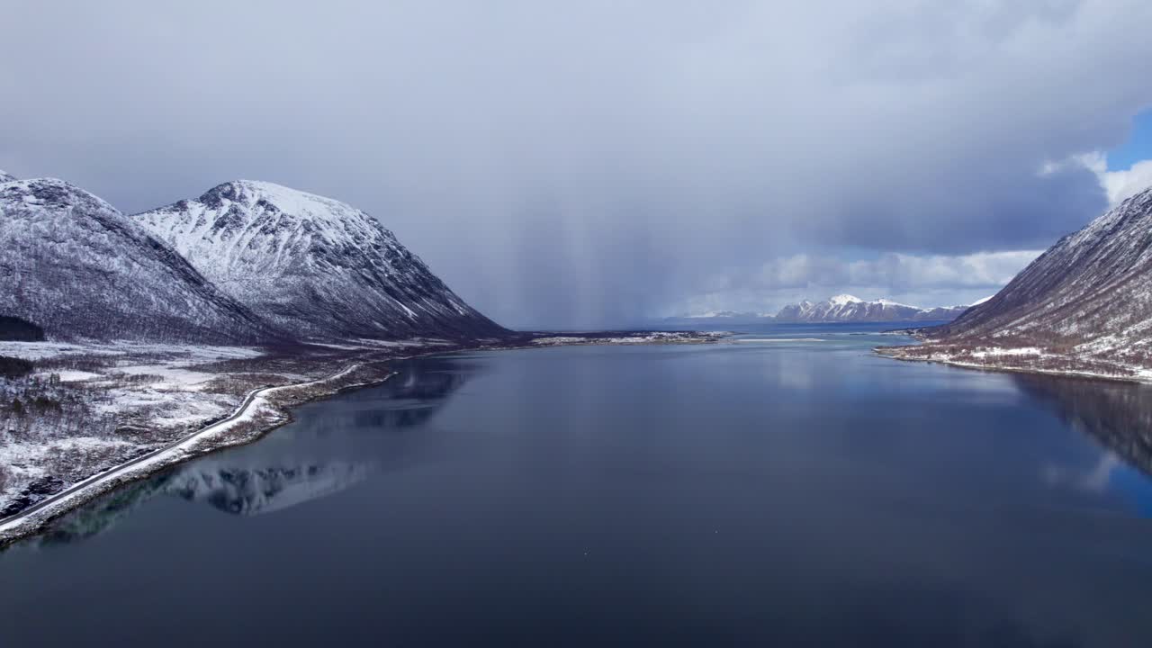 Flyfoto bakover av en storm og skyer over det stille vannet i en fjord nær Lofoten, Norge, med snø på fjellene.