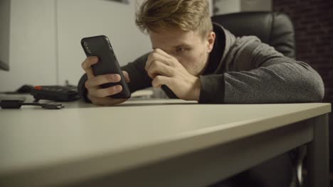 a young adult man sits at his desk as he uses his mobile phone