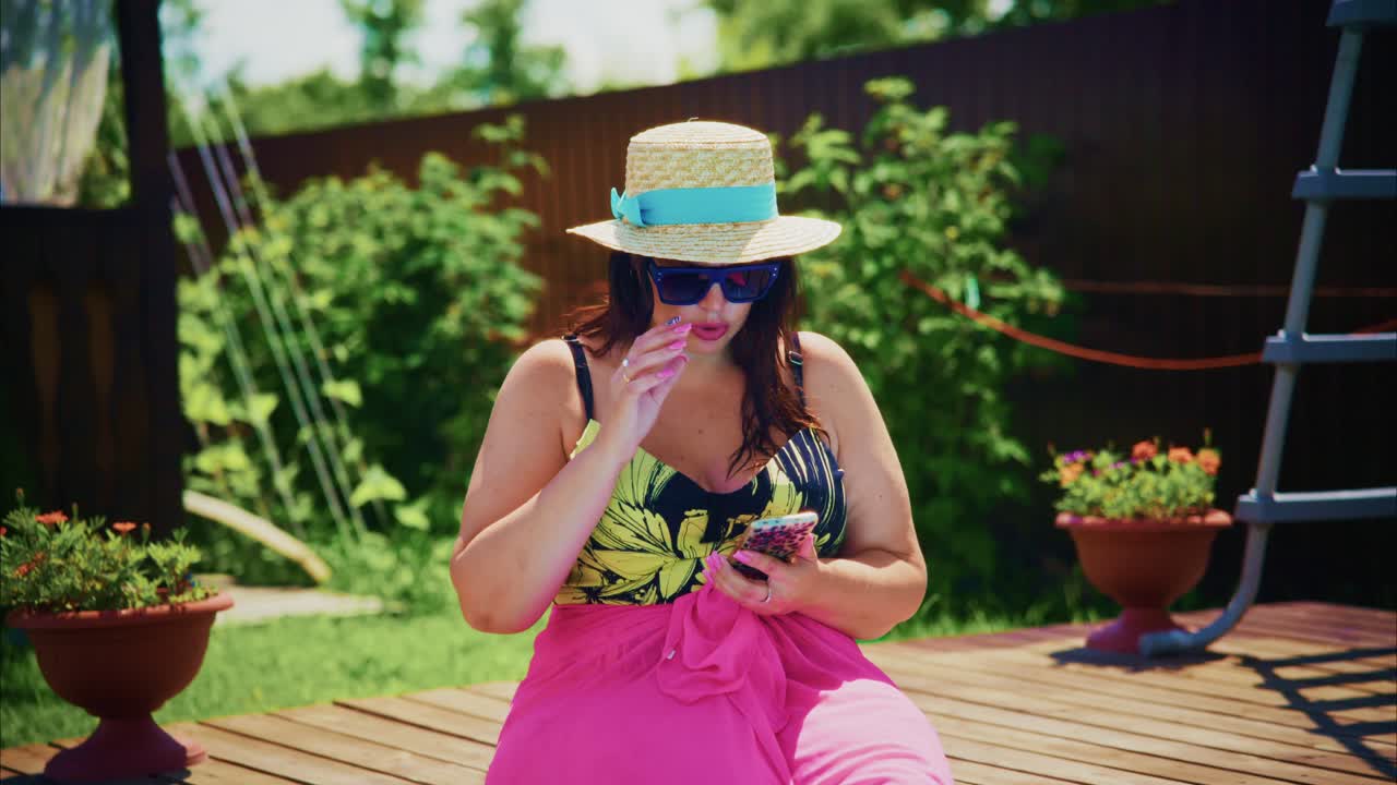 Woman in colorful swimsuit and sunhat relaxing outdoors with smartphone in a vibrant garden setting