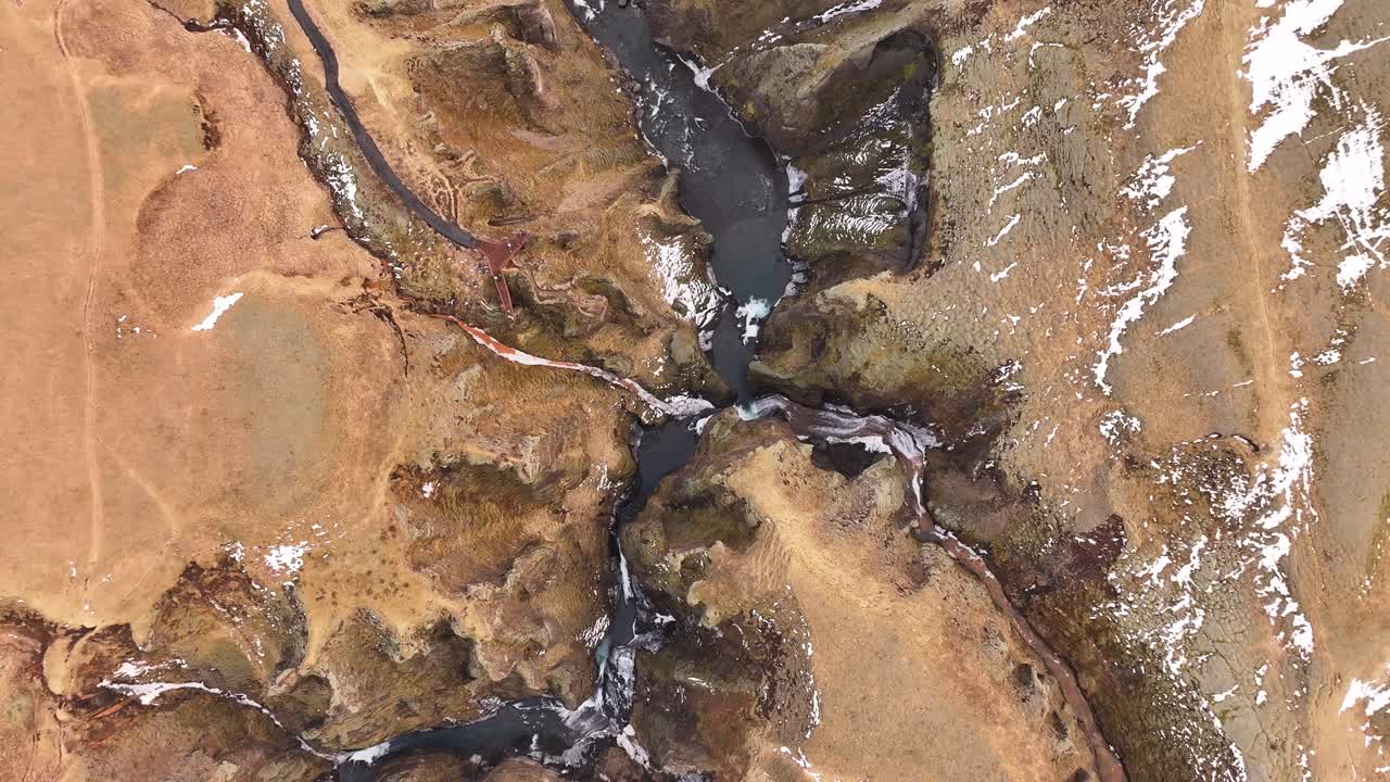 Top-down aerial view of Fjaðrárgljúfur Canyon near Kirkjubæjarklaustur, Iceland, revealing dramatic cliffs, winding river paths, and patches of lingering snow in a rugged landscape.