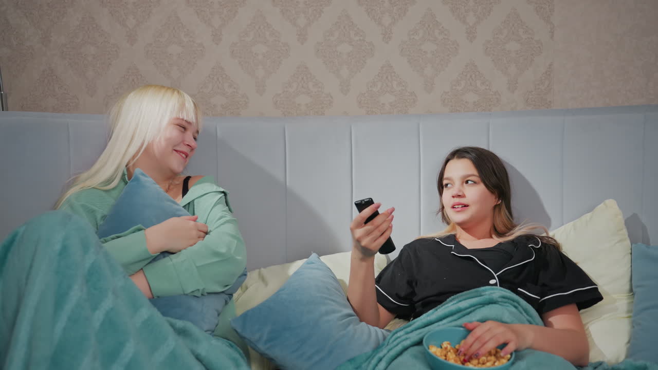 Two siblings wearing pajamas relax in bed watching TV together, one eats popcorn while holding remote, other hugs pillow, snacks are on blanket as both enjoy show
