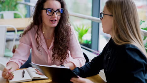 brunette and blonde long-haired ladies wearing glasses sit at table talking. female colleagues discussing job moments.