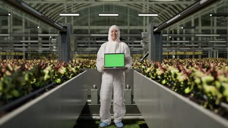 full body of asian man researcher smiling and showing green screen laptop to camera while standing in the greenhouse with smart robotic farmers