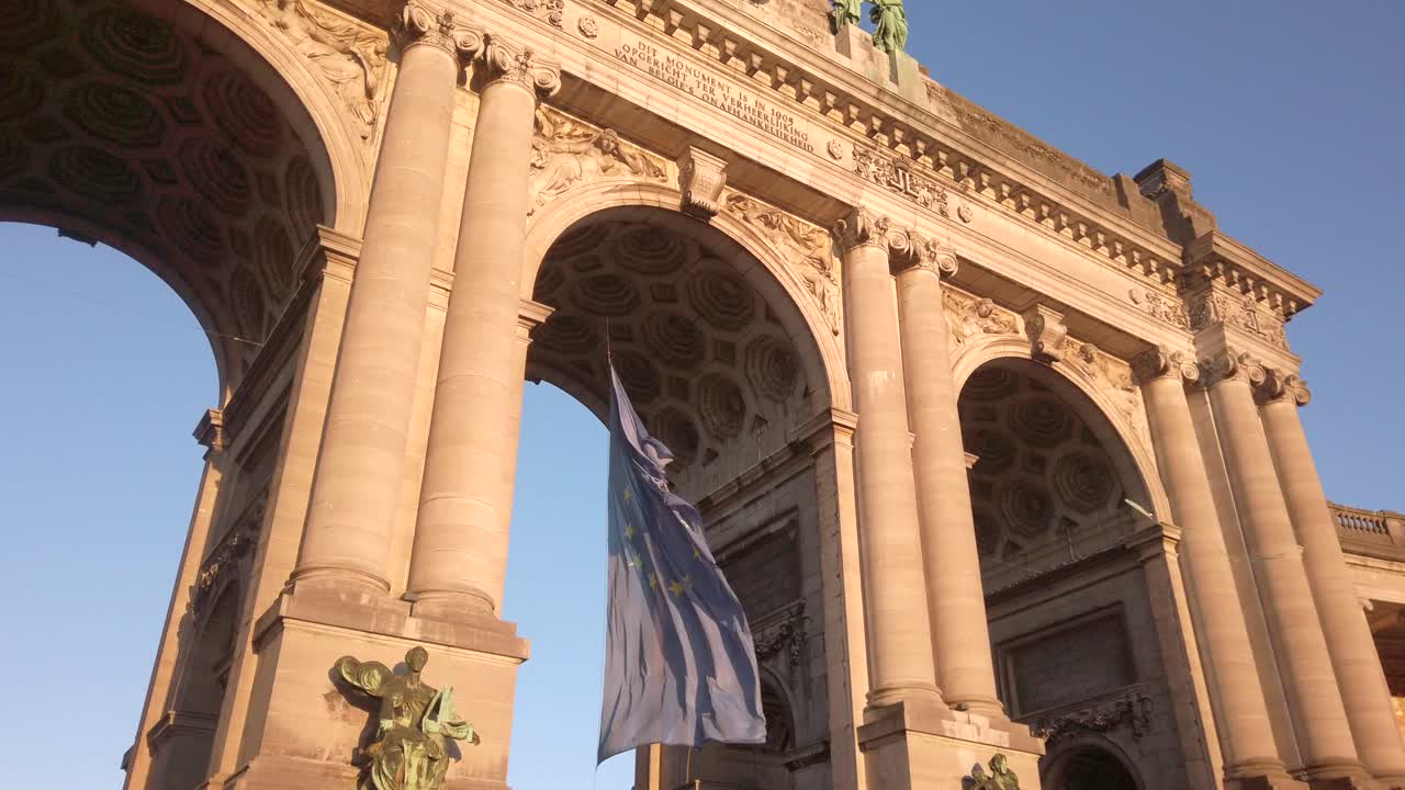 Wide-angle shot of the European Union flag waving beneath the Cinquantenaire monument in Brussels, with a playful kite soaring through the archway