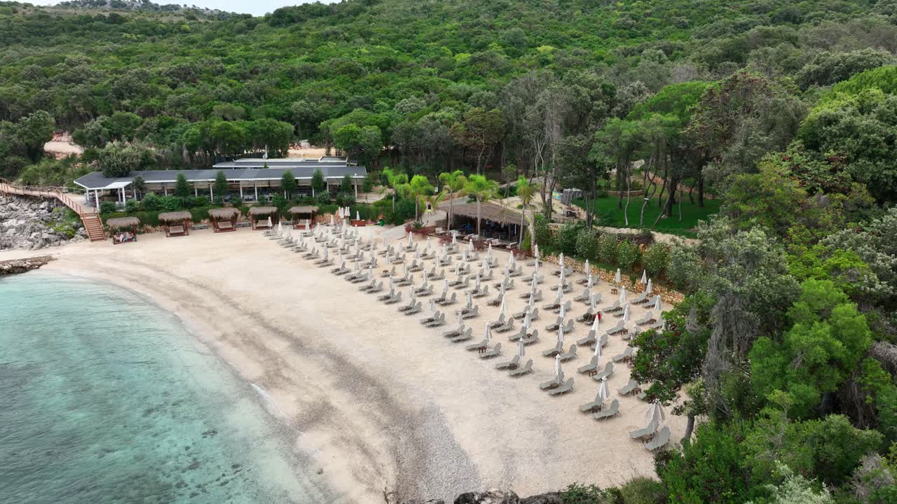 Aerial view of a stunning beach in Albania, featuring clear turquoise waters, white sand, numerous sun loungers, and lush green surroundings, perfect for tourism and relaxation.
