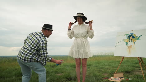 a man in a checkered shirt is seen spraying mosquito repellent on a woman's legs, who is in a stylish hat and a white dress. they stand in a grass field with a painted canvas and art supplies nearby