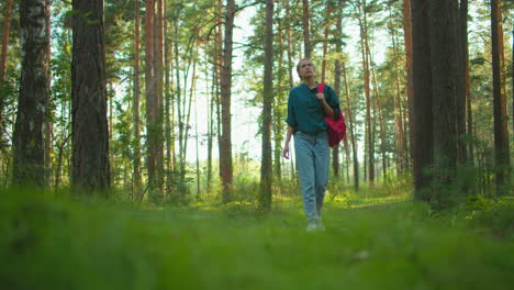 woman strolling through forest, carrying red backpack and gazing around at tall trees bathed in sunlight, surrounded by lush greenery, she explores with a calm demeanor