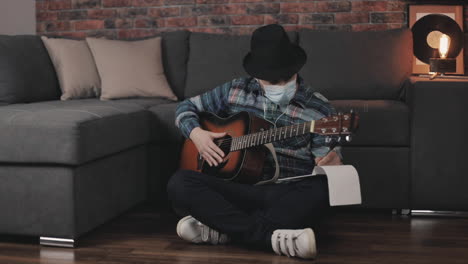 joven músico masculino con mascarilla médica creando música para guitarra, teniendo ideas, tomando notas en casa durante el encierro debido a la pandemia de covid 19