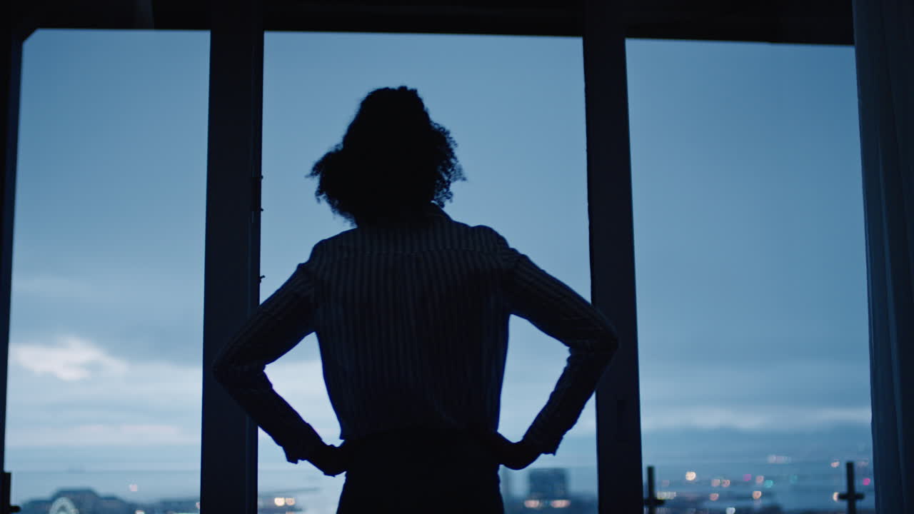 young woman looking out window enjoying view of city at night contemplating successful lifestyle planning ahead on calm urban evening