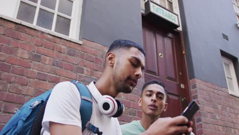 dos amigos varones de raza mixta sentados, hablando y usando un teléfono inteligente en la calle
