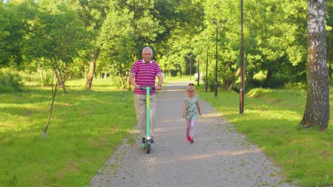 Senior-modern-man-grandfather-tourist-riding-electric-scooter-with-granddaughter-child-girl-in-park