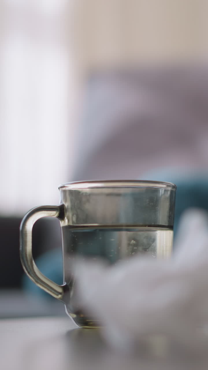 Close-up of medication, used tissue, and glass cup on table, with blurred view of sick person in background resting, showing signs of cold or flu symptoms, looking unwell