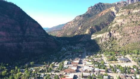 aerial drone reveal of ouray colorado mountain town, cars driving through city and houses surrounded by rocky mountain cliffs and pine tree forest