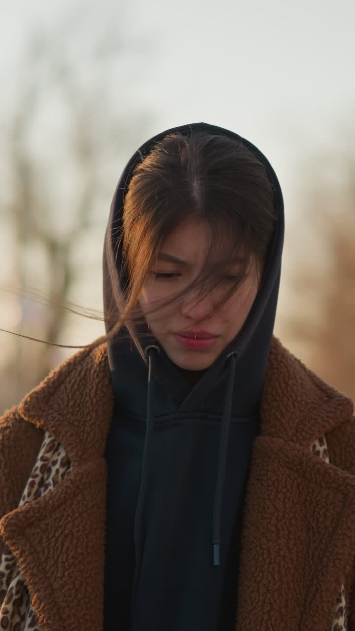 Front shot of a sad girl walking through a lonely park at sunset, wearing a brown coat and a hoodie. melancholic mood with the girl's downcast expression and the serene, empty park setting