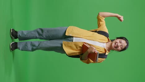 full body of asian woman student with a backpack and some books smiling and flexing her bicep while standing in the green screen background studio