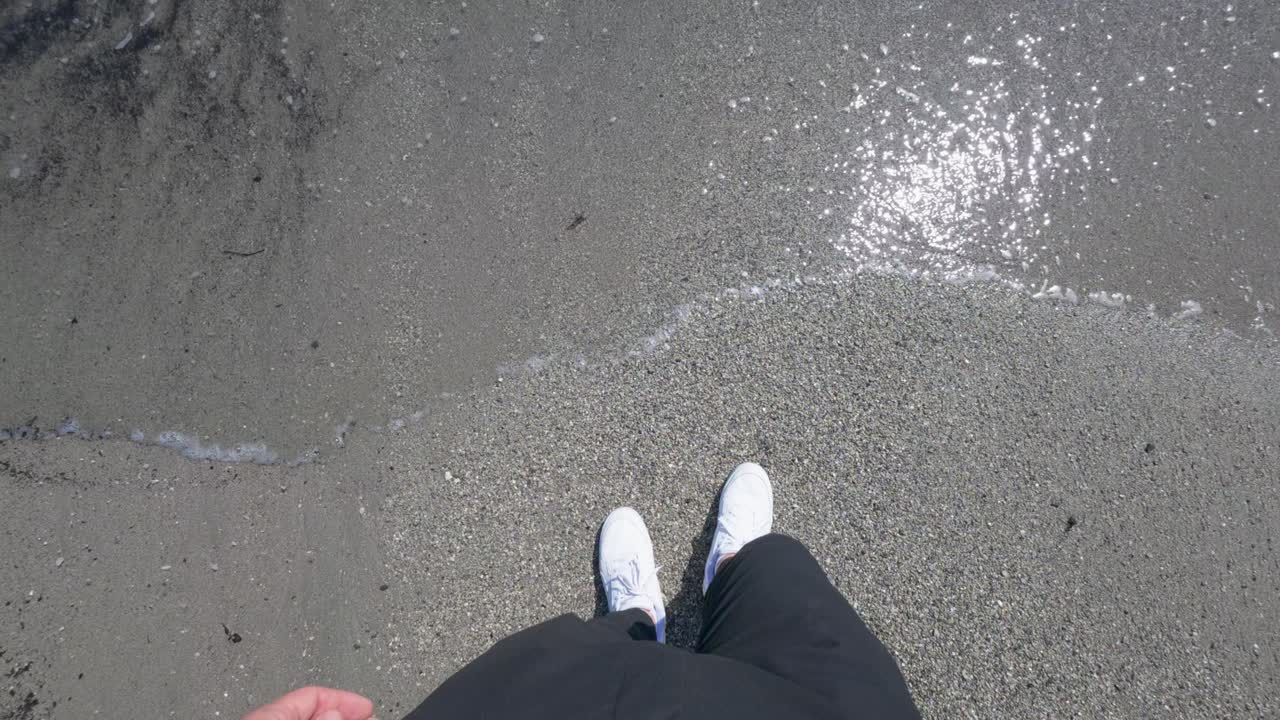 Beyaz spor ayakkabılı bir kişinin ıslak kumda durduğu, güneş ışığının sahilde yansıdığı ve sakin dalgaların kıyıya vurduğu yukarıdan bakış açısı çekimi. Gerçek zamanlı, doğal gün ışığı, elde kamera hareketi.
