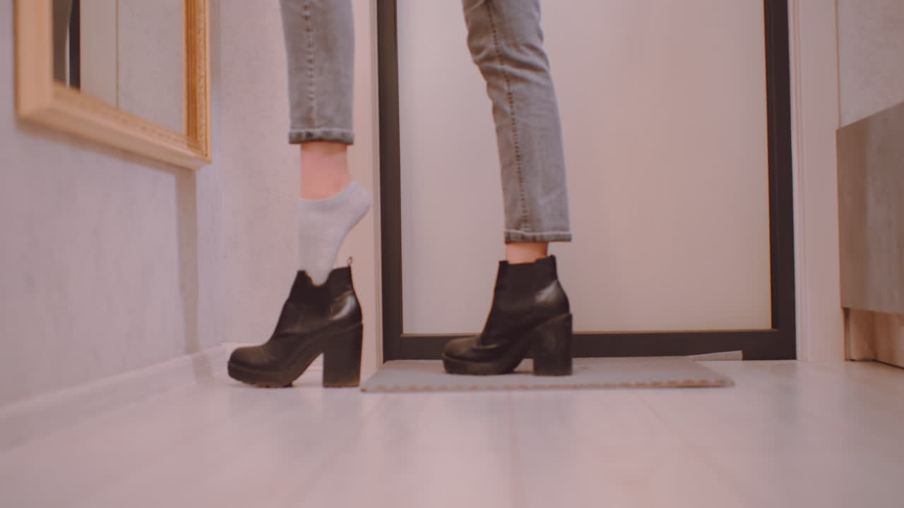 Lower angle close view of young woman in jeans and sweater putting on black heeled shoe while one foot still in sock, preparing to go out in modern hallway