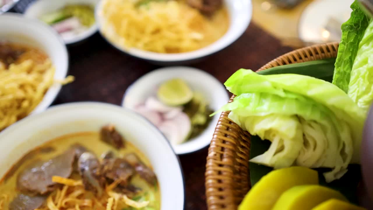 A vibrant spread of Northern Thai dishes including beef curry noodles, roasted green chili dip, fresh vegetables, boiled egg, and steamed rice, shot in natural light with shallow depth of field and smooth camera movement