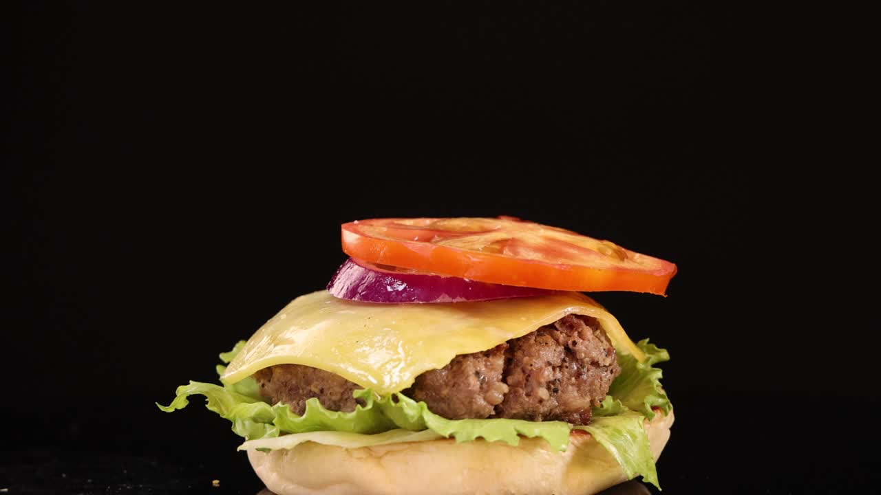 A sesame seed bun top falls onto a stacked beef burger with lettuce, cheese, tomato, and onion, shot in slow motion against a black background with studio lighting