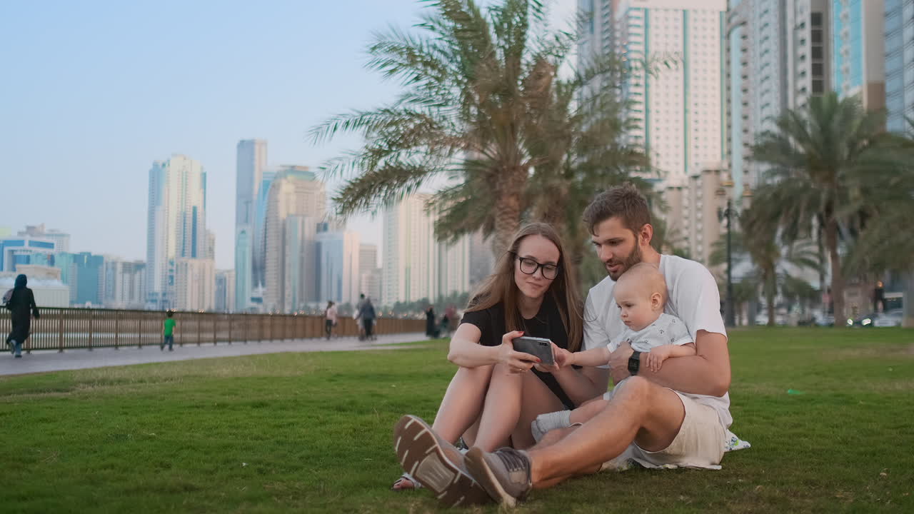 Happy family with one children sitting together on grass in park and taking a selfie. With smartphone.