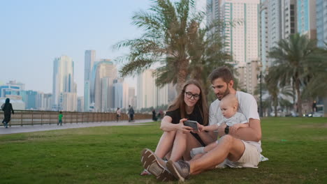 Happy-family-with-one-children-sitting-together-on-grass-in-park-and-taking-a-selfie.-With-smartphone.