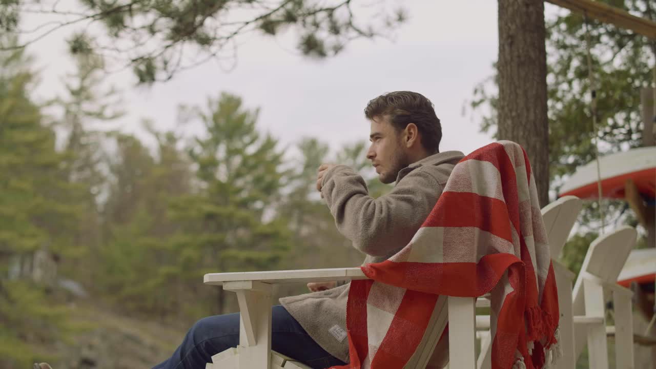 hombre relajándose en un acogedor entorno al aire libre, bebiendo una bebida mientras está envuelto en una manta y rodeado de naturaleza
