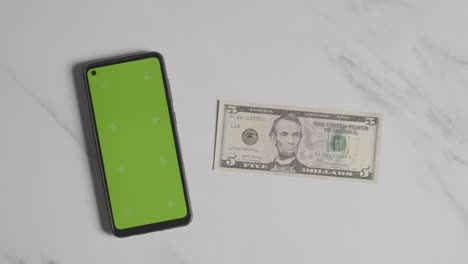 overhead currency shot of hand putting down us 5 dollar bill next to green screen mobile phone