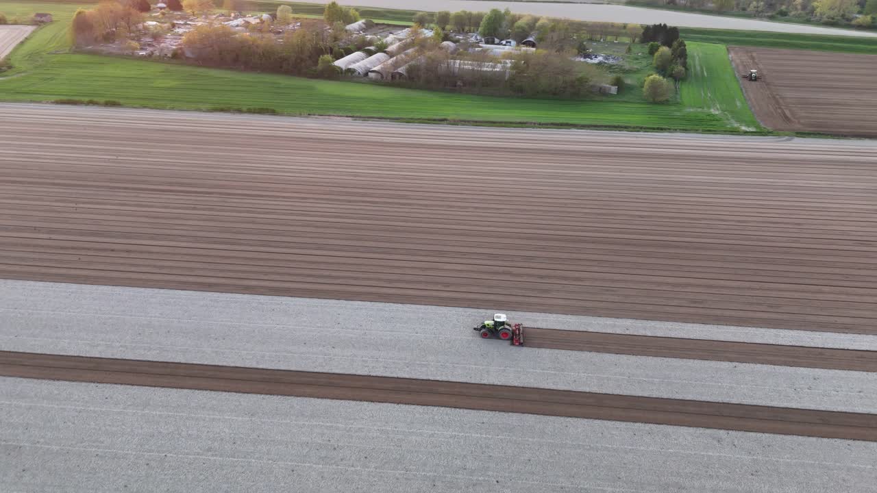 Slow orbiting drone captures tractor pulling leveling harrow as it carves deep brown seed rows through bleached soil, set against green edge of farmyard and distant fields under soft sunset light