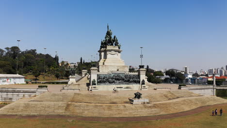 Toma-Aérea-Acercándose-Al-Monumento-A-La-Independencia-Pasando-Por-El-Fuego-De-La-Libertad-En-Primer-Plano-Con-El-Museo-Ipiranga-En-El-Fondo-De-La-Escena