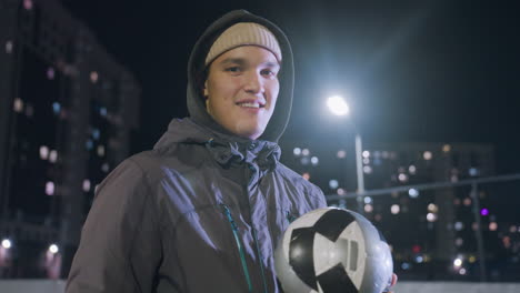 joven con capucha sonriendo a la cámara mientras juega a golpear el fútbol en su mano durante la noche en el campo deportivo urbano rodeado de edificios residenciales iluminados y luces de calle brillantes