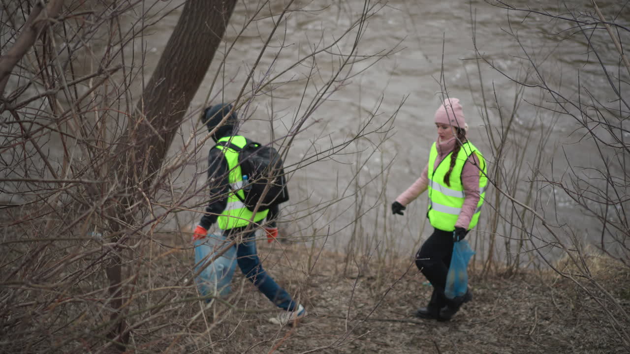Two volunteers in reflective vests carrying blue bags filled with collected trash walking along riverbank through leafless branches during environmental cleanup on cold overcast day