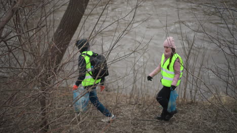 two volunteers in reflective vests carrying blue bags filled with collected trash walking along riverbank through leafless branches during environmental cleanup on cold overcast day