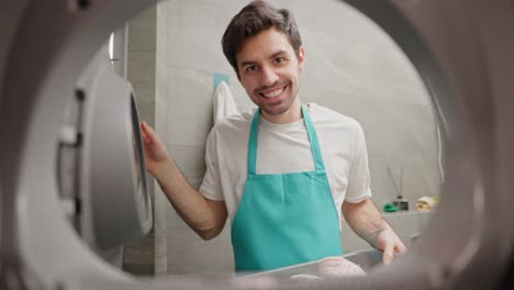 Portrait-of-a-creepy-brunette-janitor-a-man-in-a-white-T-shirt-and-blue-apron-slowly-closes-the-washing-machine-door-and-looks-inside-with-open-eyes-and-a-creepy-smile-while-cleaning-in-a-modern-apartment