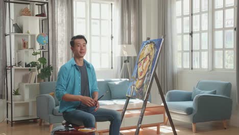 asian artist man sitting on round wooden chair without backrest holding paintbrush mixed colour wipe the sweat before crossing his arms and smiling after finish painting a girl on the canvas