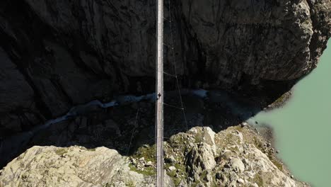 a static, symmetrical shot of a solo, young man is walking alone by himself across trifthutte in switzerland on a suspension bridge, in perfect center and middle above a small river or stream