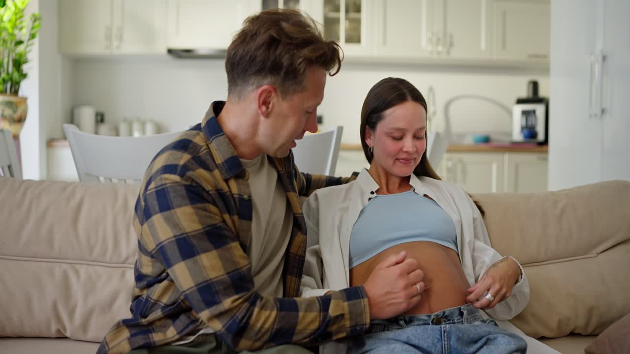 Happy man in a plaid shirt communicates with his pregnant wife and strokes her chiffon at home on the sofa in a modern apartment