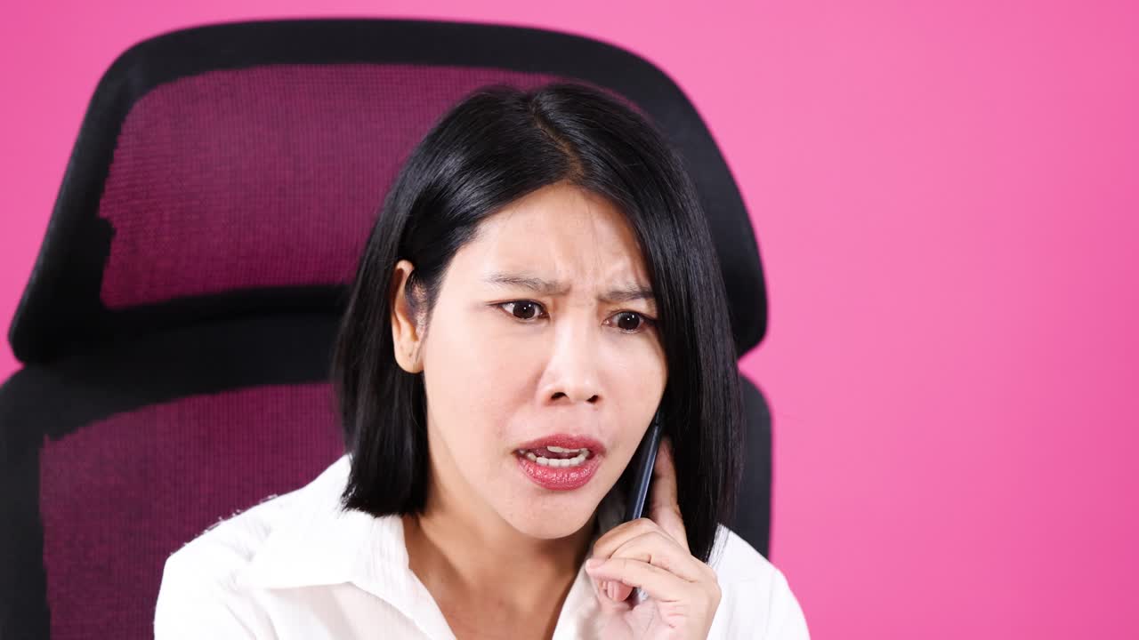 Asian woman in white shirt expresses surprise and concern on phone, seated against pink background