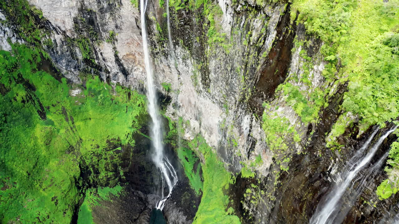 Veduta aerea con drone di più strette cascate che scendono da ripide scogliere verdi, circondate da una fitta vegetazione in un paesaggio montano tropicale.