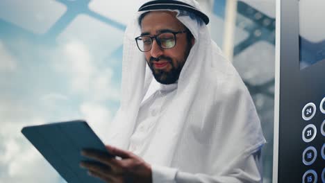 successful muslim businessman in traditional white kandura riding glass elevator to office in modern business center. man using tablet computer. saudi, emirati, arab businessman concept.