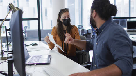 Verschiedene-Männliche-Und-Weibliche-Kollegen-Mit-Gesichtsmasken-Sitzen-Am-Schreibtisch-Und-Reden-Und-Stoßen-Mit-Dem-Ellenbogen-Aneinander