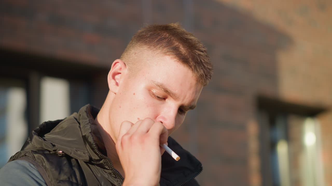 close up of young man in sunlight holding cigarette near lips then deciding not to smoke and throwing cigarette away, standing in front of brick wall with serious expression and soft natural lighting
