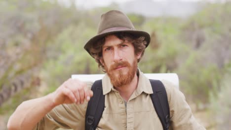 Portrait-of-serious-bearded-caucasian-male-survivalist-trekking-through-wilderness,-raising-hat