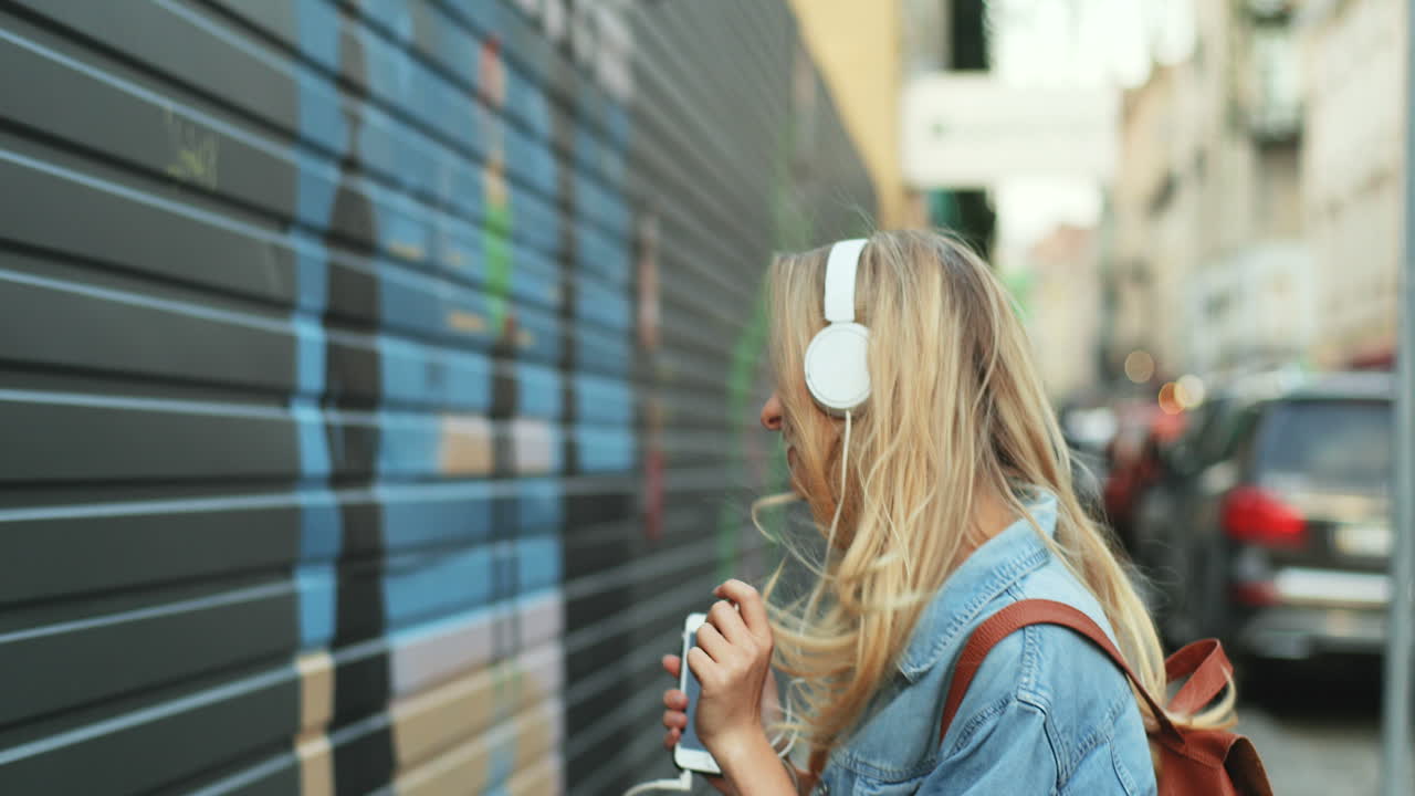 Caucasian blonde young woman listening to music with headphones and dancing in the street