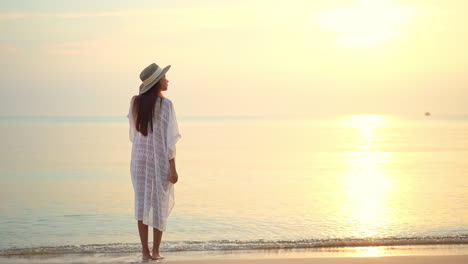 back view of barefoot woman with hat and dressed in white on beach at sunset