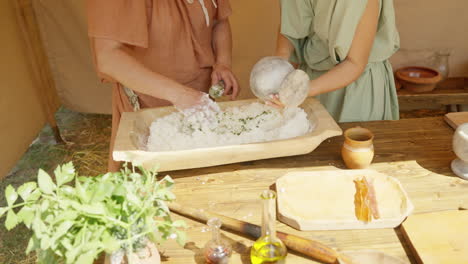 a slow pull-out shot shows two people in ancient roman costumes preparing food. they mix flour and herbs in a wooden bowl to make bread, demonstrating historical cooking