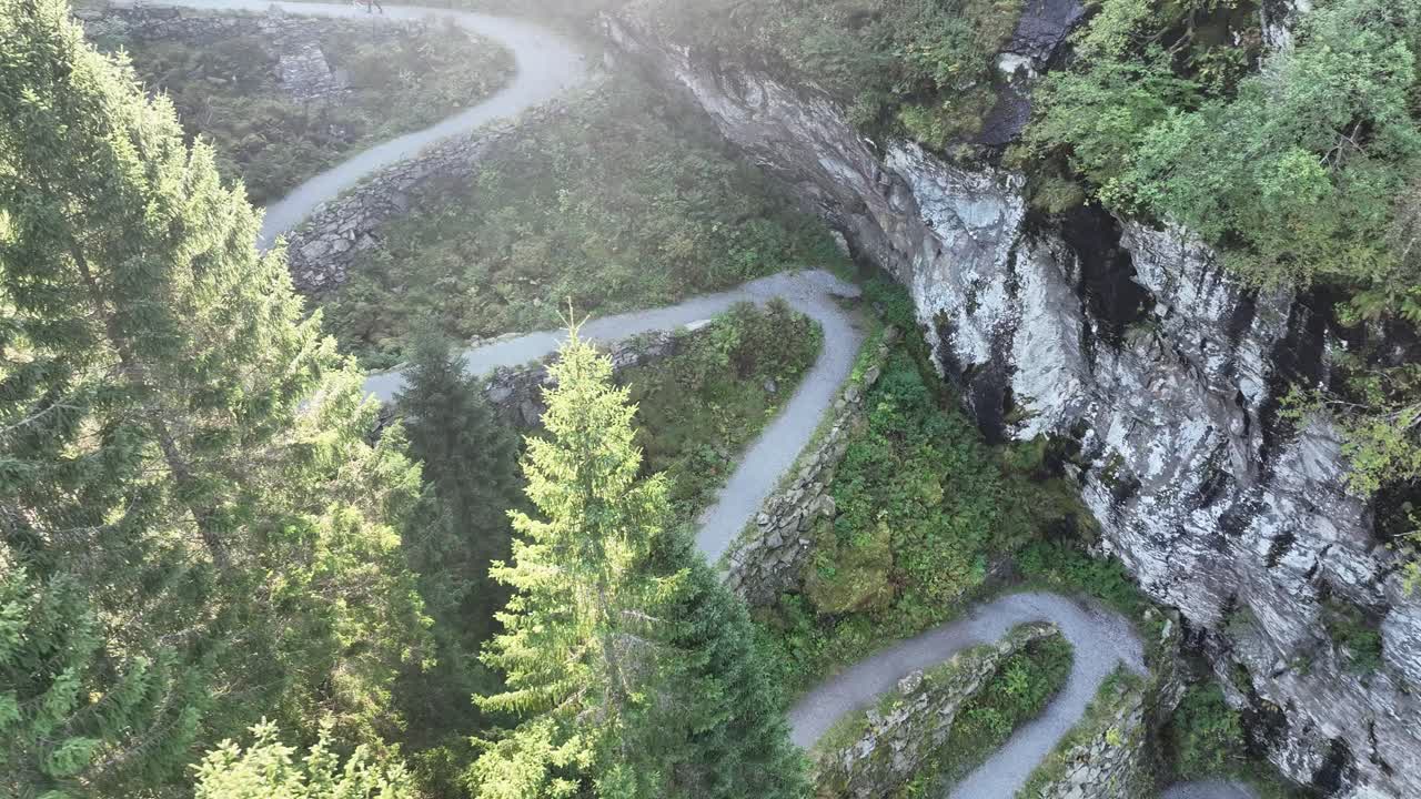High angle aerial of winding road through spruce trees with sunrays. Two people walk up the road, slow parallax motion