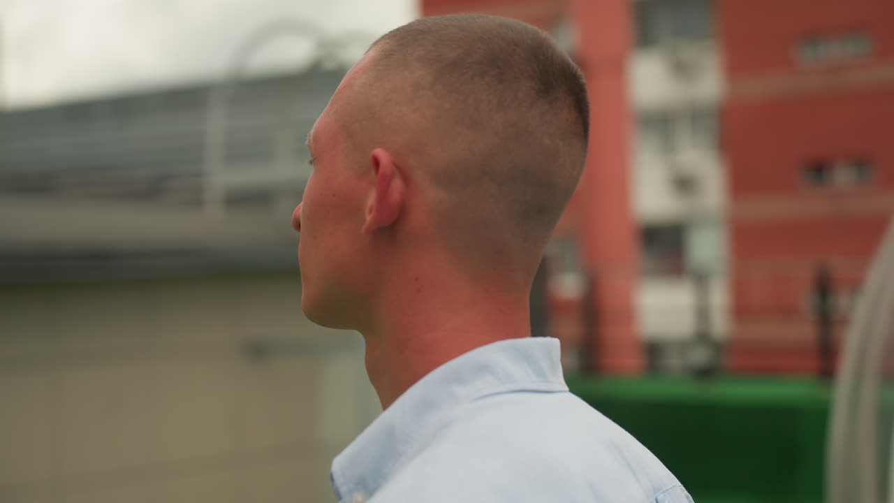Uomo su un tetto che osserva lo skyline urbano, ritratto riflessivo che cattura la luce della sera, vento che scompiglia i capelli corti, camicia casual ed espressione discreta, ringhiera sul tetto ed edifici distanti che incorniciano la scena