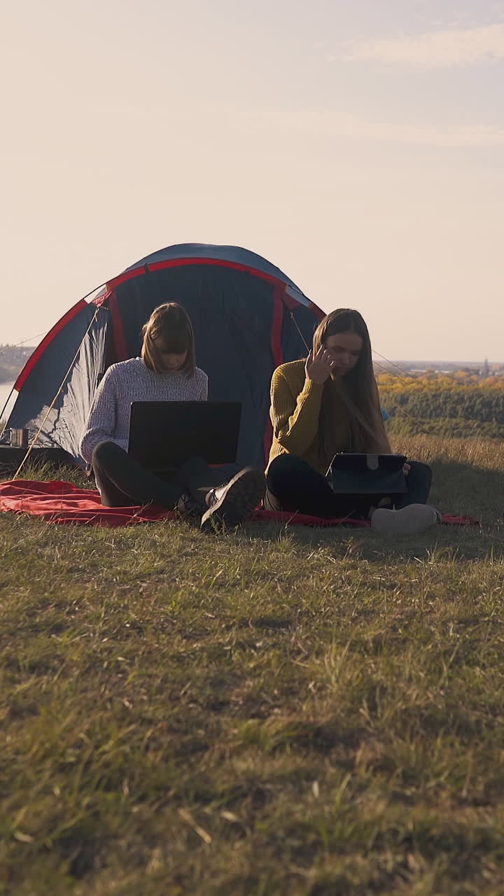 pretty girls with modern tablet and laptop sit near blue tent in camp on steep river bank in warm autumn morning